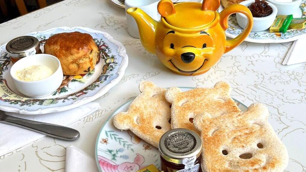Petit-déjeuner sur une table avec une théière en forme de personnage de dessin animé, deux toasts en forme de tête d’ours, des scones, de la confiture et du beurre.