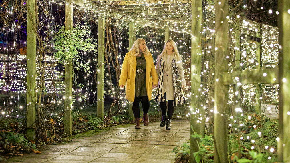 Two women walking together through a lit walkway covered in plants.