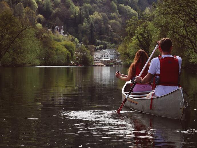 Due persone remano in una canoa su un fiume calmo circondato da alberi, con case sullo sfondo.