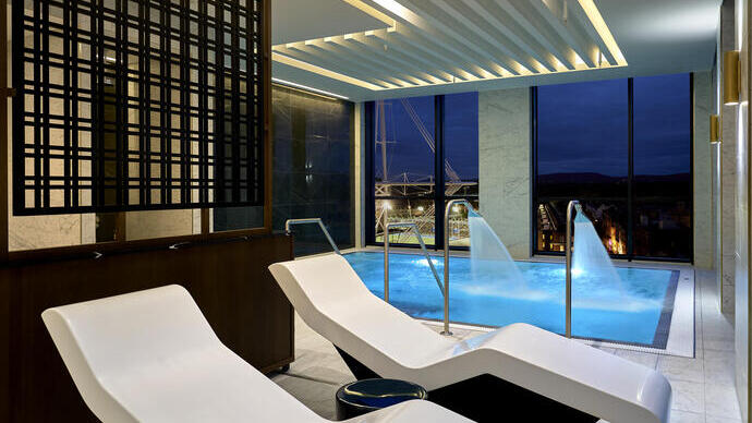 Modern indoor spa with lounge chairs, a pool with water jets, and large windows overlooking a city at night.