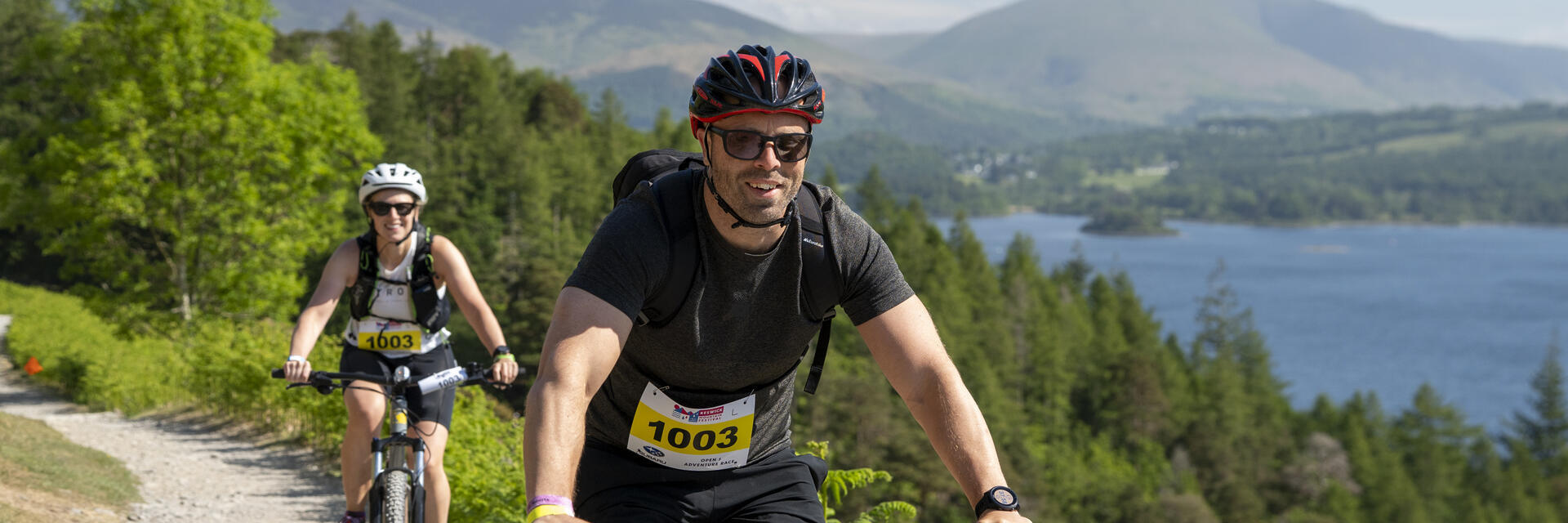Two cyclists ride on a scenic trail beside a lake, surrounded by green hills and distant mountains under a blue sky.