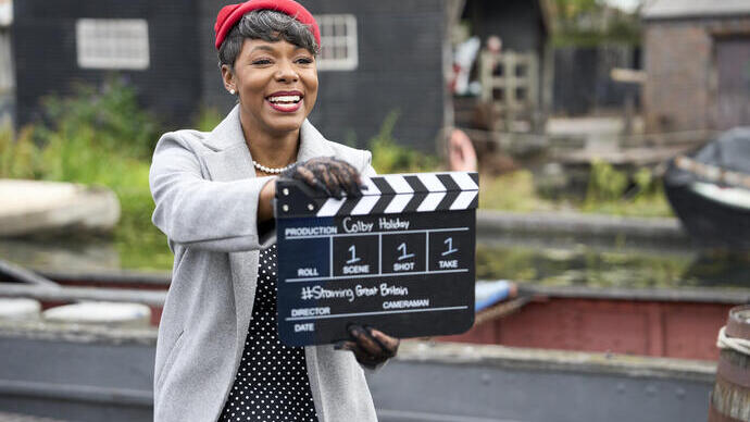 Lächelnde Frau hält eine Filmklappe an einem Drehort im Freien.