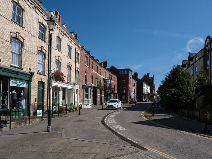 Street scene with traditional brick buildings, shops, parked cars, and a clear blue sky on a sunny day.