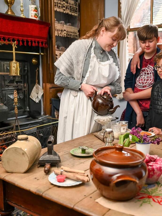 A group observing a kitchen scene from the past with a lady pouring tea at the table inside a living museum.