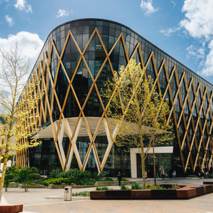 Modern building with gold lattice exterior, large glass windows, landscaped plaza, trees, and blue sky with clouds.