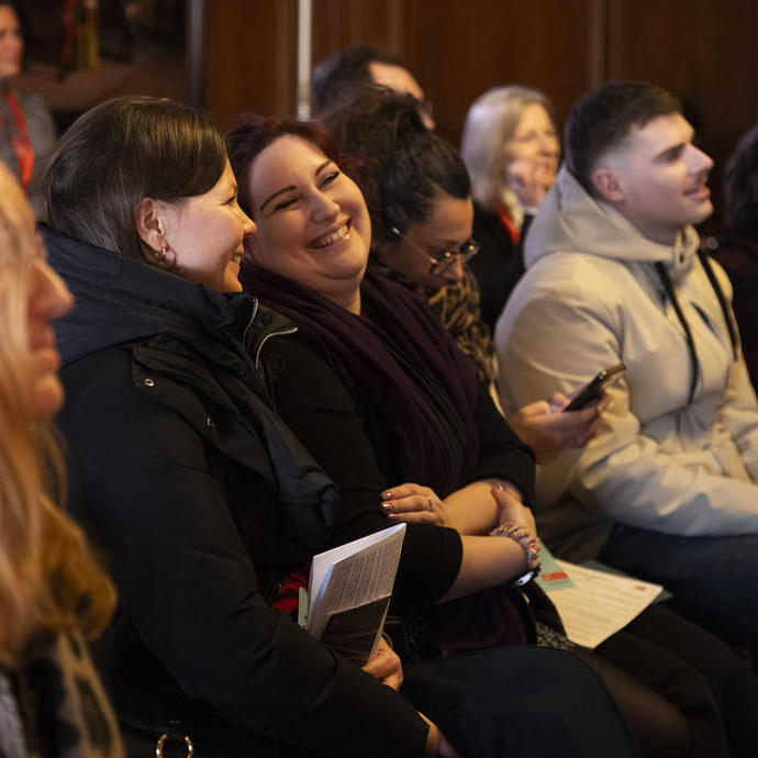 A group of people sitting indoors at an event, some smiling and laughing, with papers and phones in their hands.