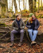 Un homme et une femme en tenue de randonnée se sourient, assis sur des racines d'arbres dans une forêt.