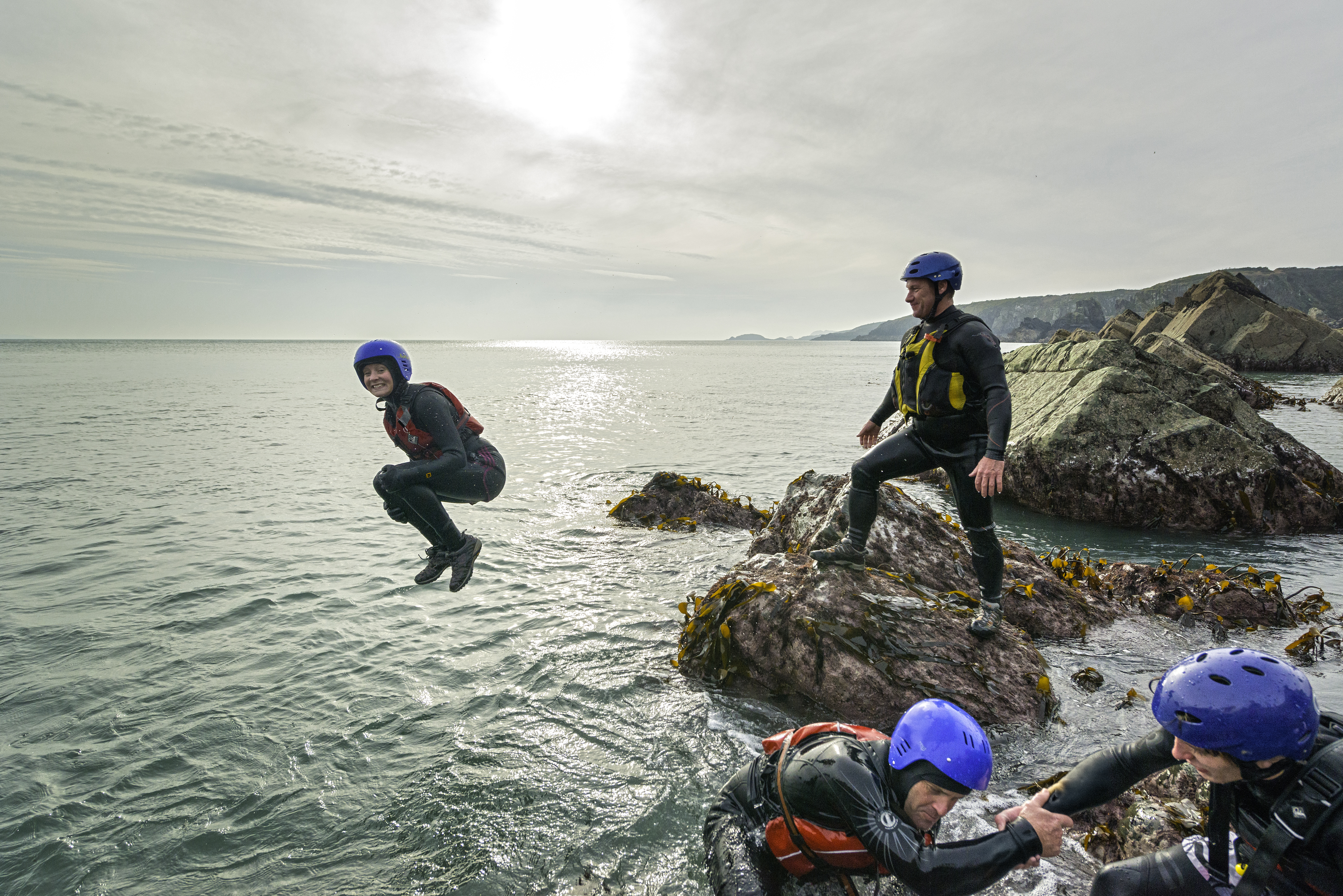 People in wetsuits and helmets coasteering by the sea, jumping and climbing on rocks under a cloudy sky, with scenic coastline in the background.