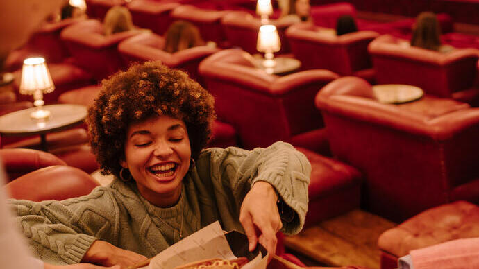 A woman taking a hot dog from a food tray being handed to her by a man inside a retro cinema.