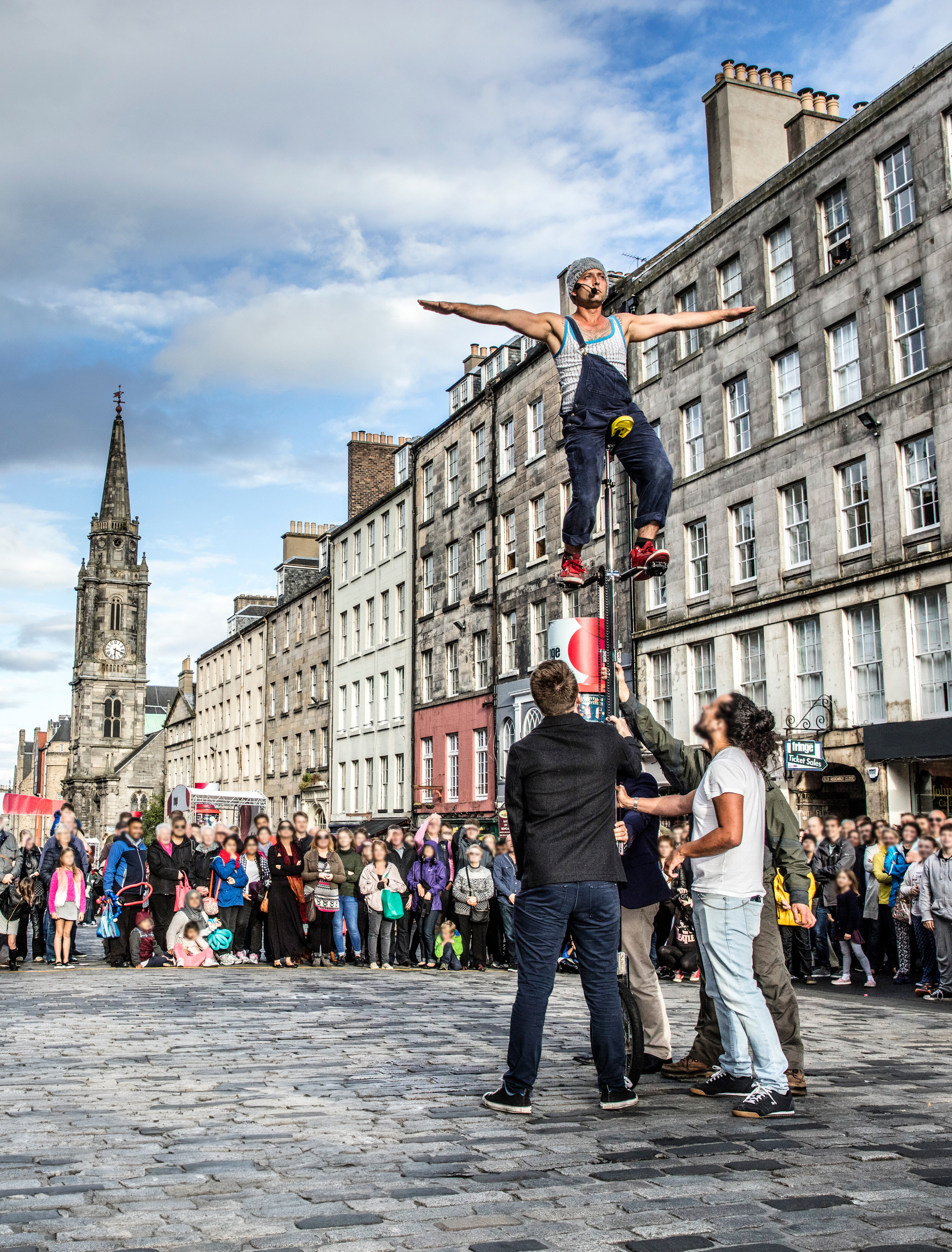 Man balancing high up on a unicycle and being held by helpers as a crowd stand watching in the street