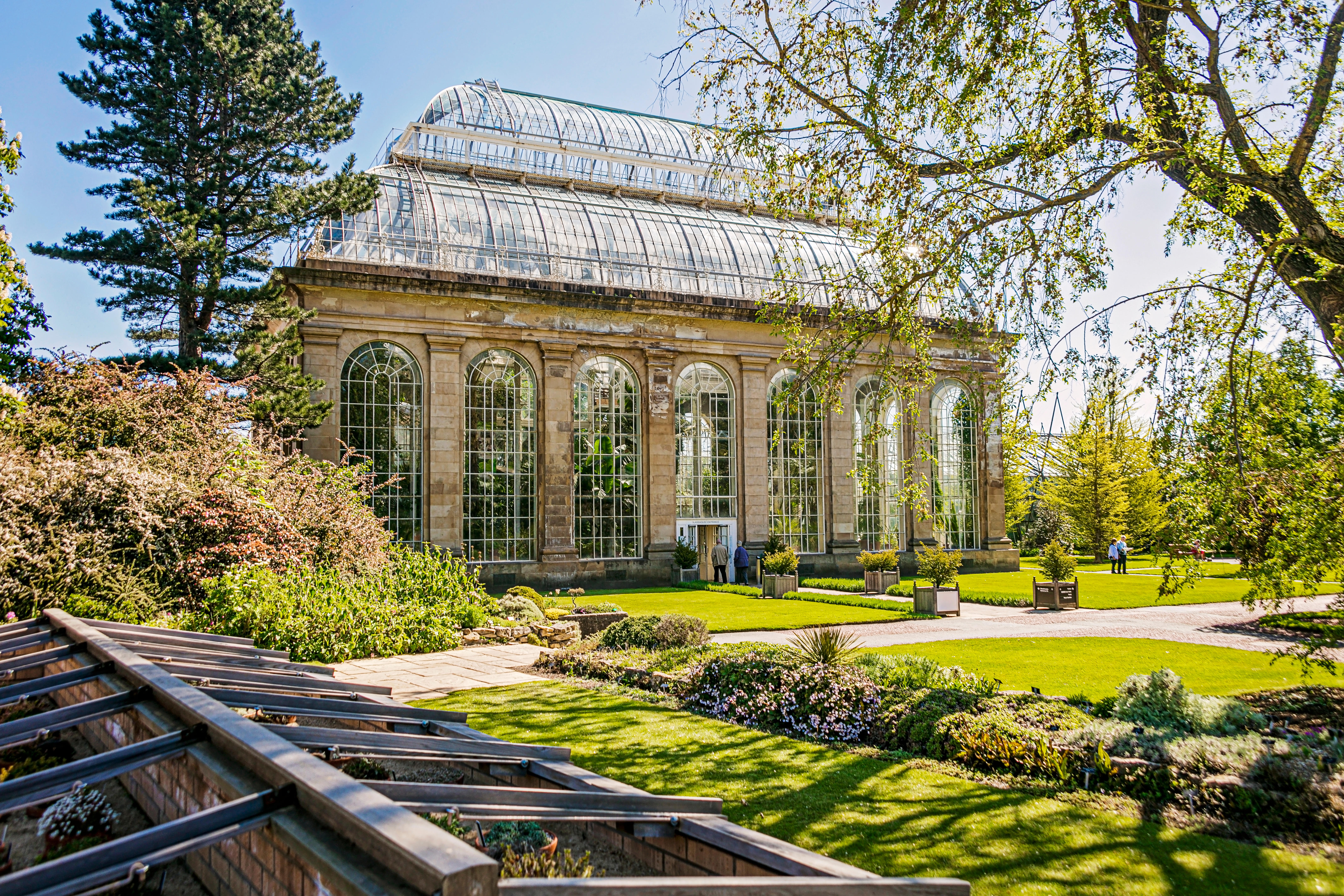 Large glasshouse with arched windows in a botanical garden, surrounded by green lawns, trees, and landscaped flower beds on a sunny day.