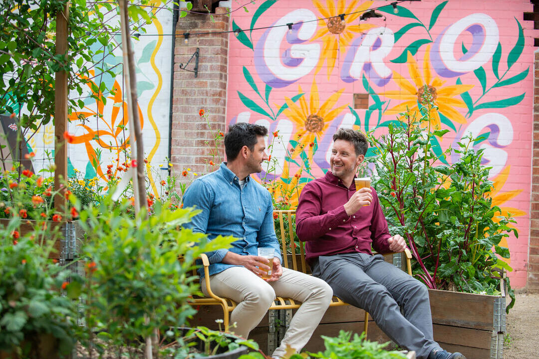 Two men sat on a bench in a garden with a drink