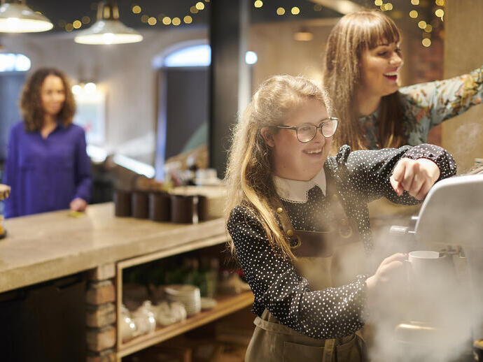 A young woman working the coffee machine with colleague assisting in cafe
