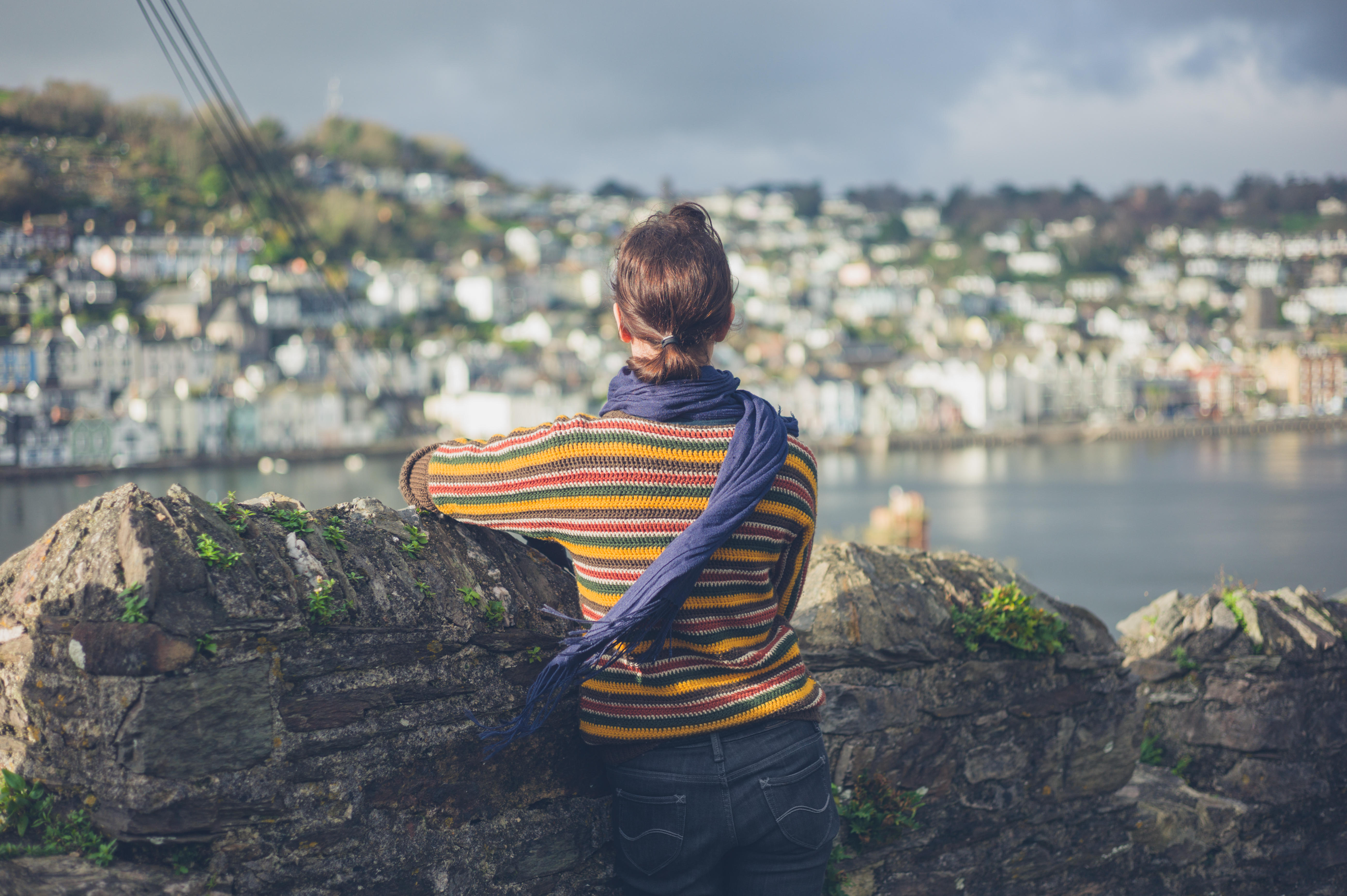 A young woman is standing by a stone wall in a village and is looking at the boats moored in the estuary