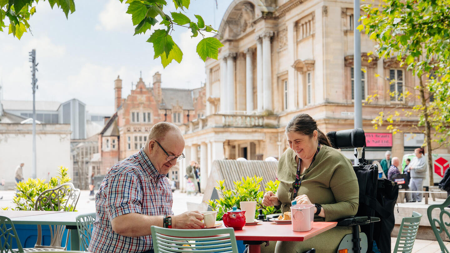 Two people sit at an outdoor café enjoying food and drinks, with a domed historic building and leafy trees in the background.