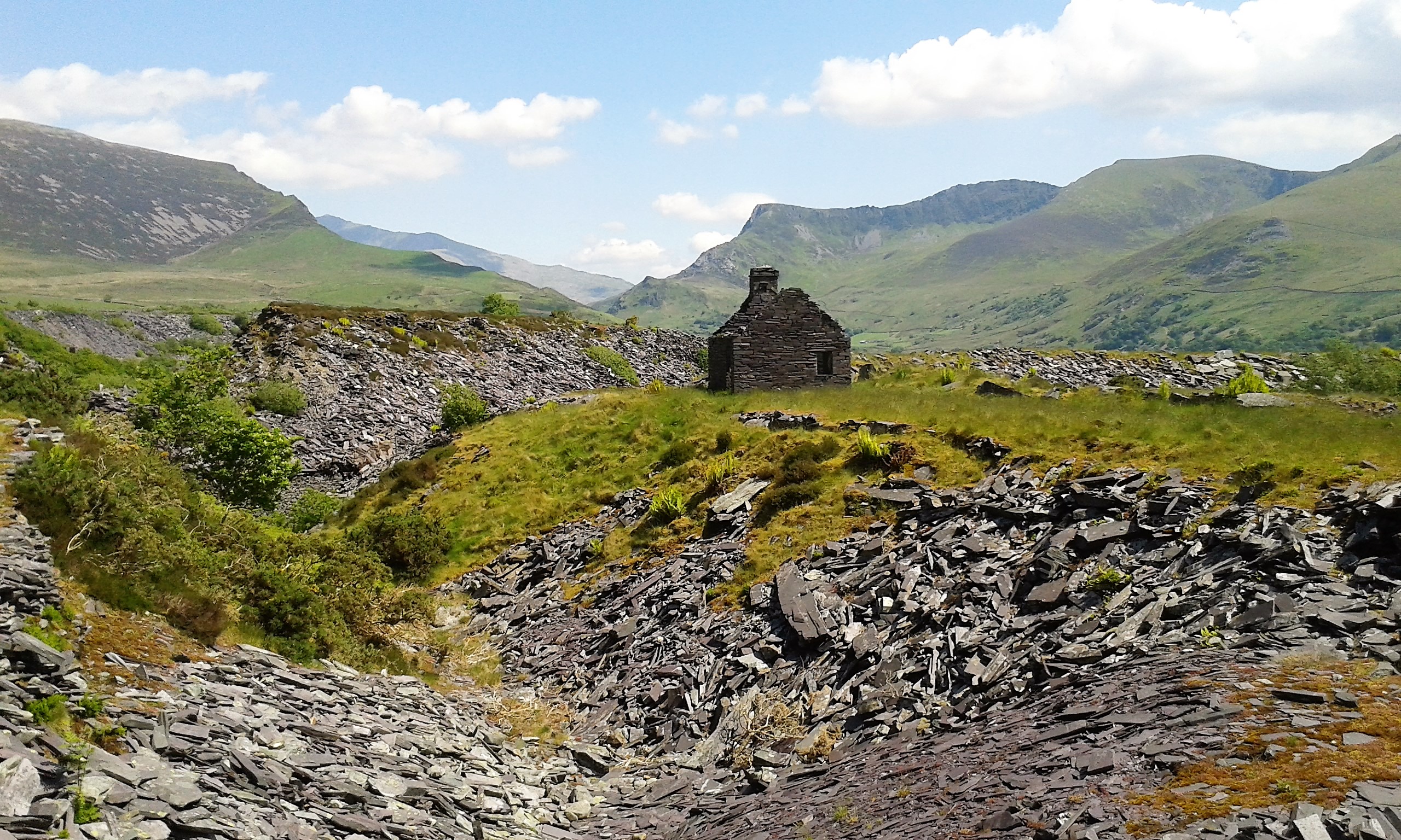 Ruined stone building on grassy hill, surrounded by slate and scattered rocks, with mountains and blue sky in the background.