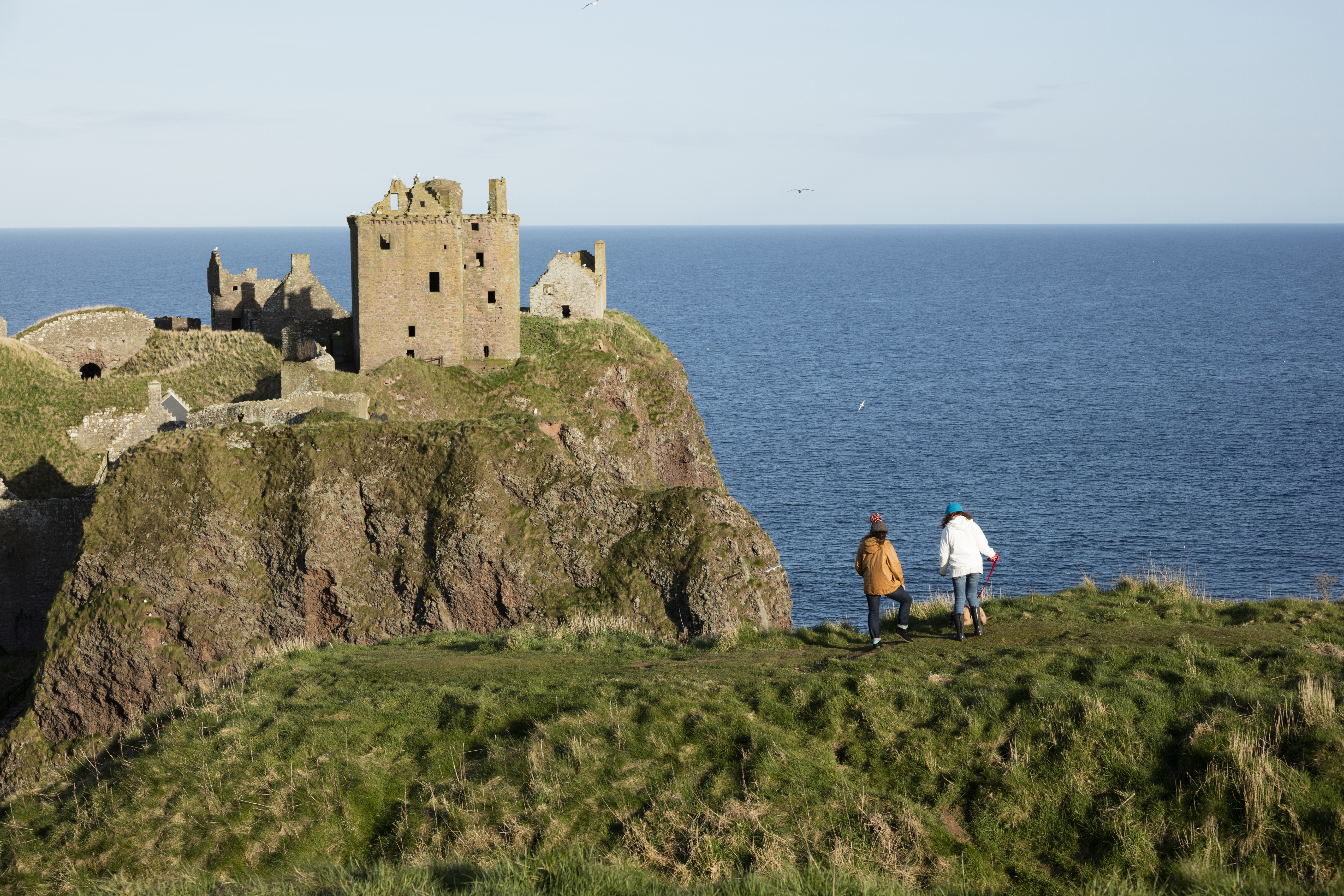 Dos mujeres de pie en lo alto de un acantilado, cerca de un castillo y del mar