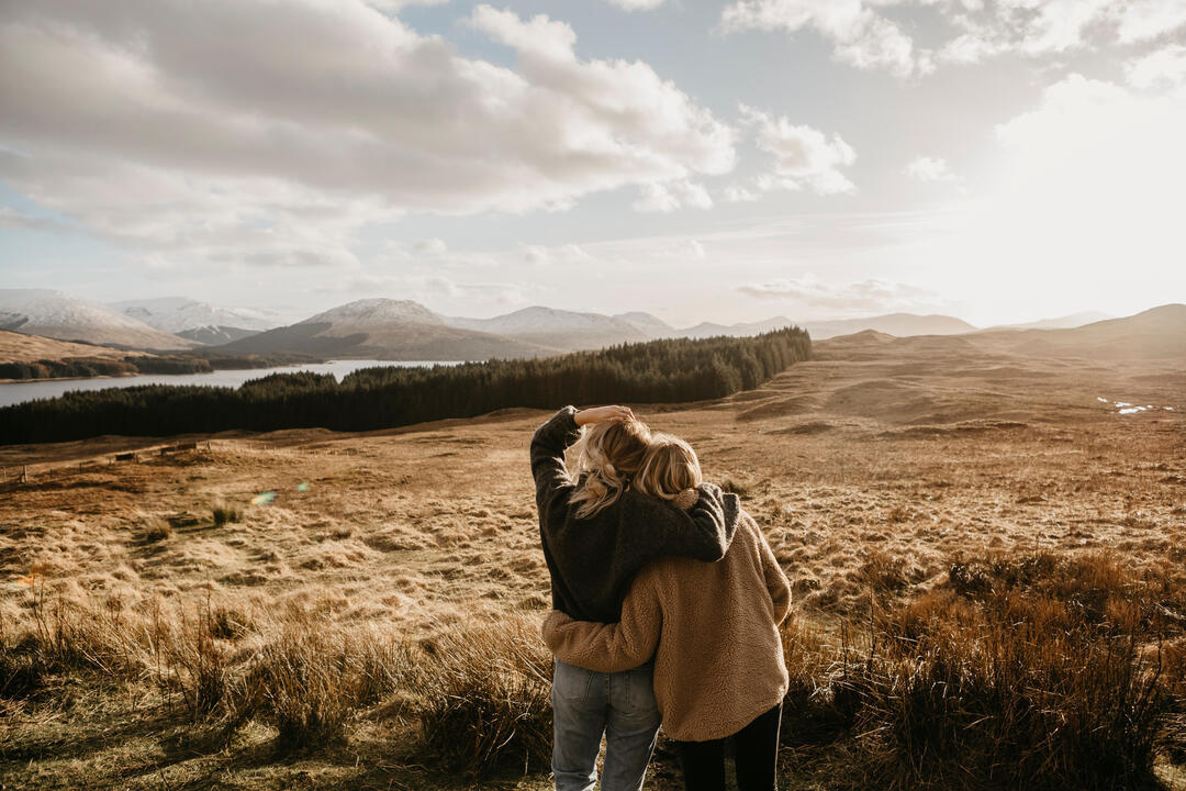 Two friends hugging looking over the valley towards a lake and mountains