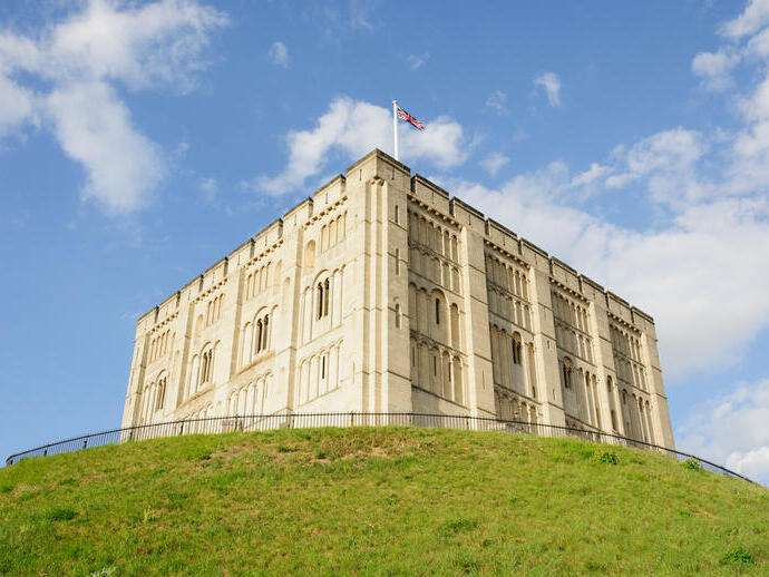 A large stone castle sits atop a grassy hill under a blue sky with scattered clouds, a flag flying above the building.