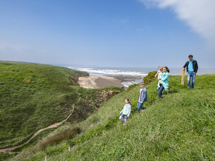 A family is walking along a grassy coastal cliff with a sandy beach and waves in the background on a clear day.