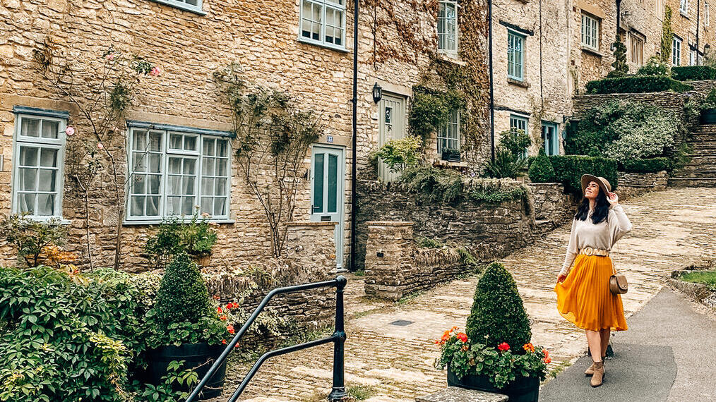 A woman in a hat walks down a steep pebbled footpath in a quaint village.