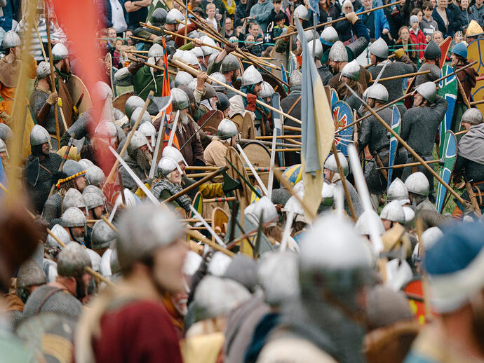 Large crowd of people in historical costumes reenact a medieval battle with shields and spears, watched by an audience.