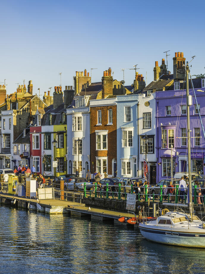 People walking up and down the water front of a busy seaside town lined with colourful terraced houses and boats anchored in the harbour
