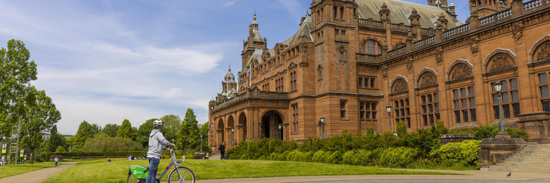Person mit Fahrrad vor einem historischen roten Backsteingebäude bei blauem Himmel an einem sonnigen Tag.