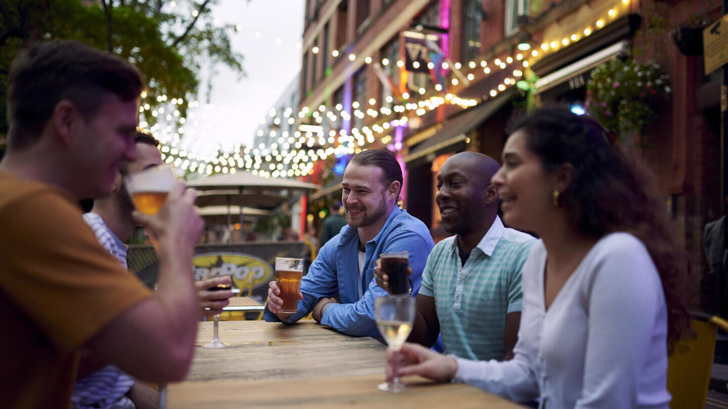 Amigos tomando una copa sentados en una mesa exterior de un bar con guirnaldas de luces colgando detrás