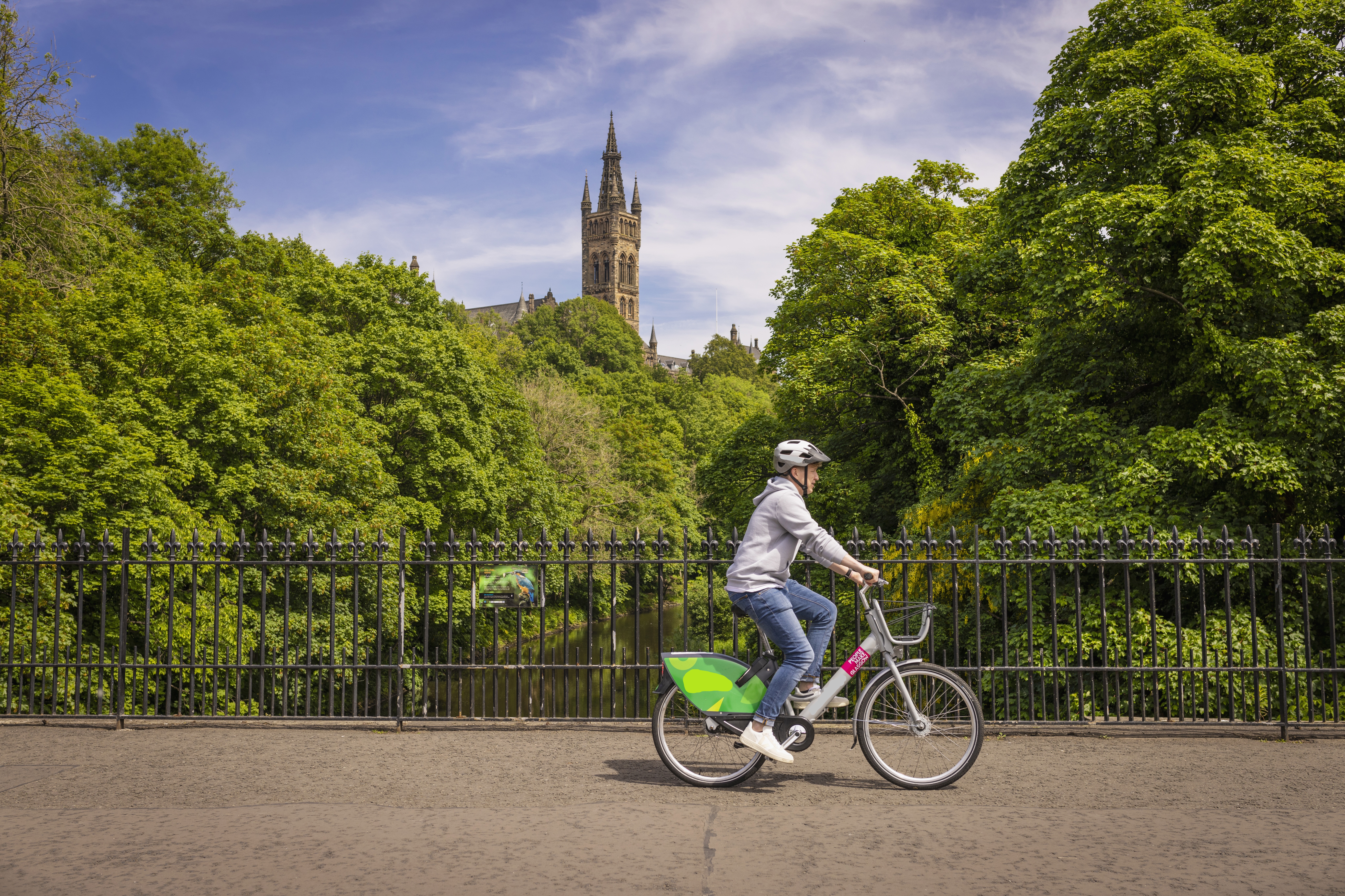 A Young Person Cycling An Ovo E-bike Over The River Kelvin in Glasgow’s popular Westend with a university and park in the background.