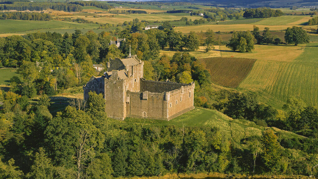 Esterno di un castello medievale in parte in rovina su una piccola collina che si affaccia su campi e alberi in autunno.