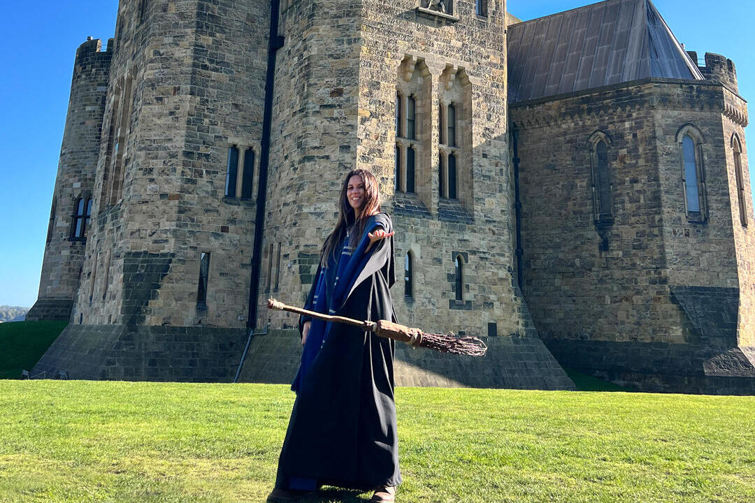 A woman poses with a cloak and broomstick in front of an heritage castle