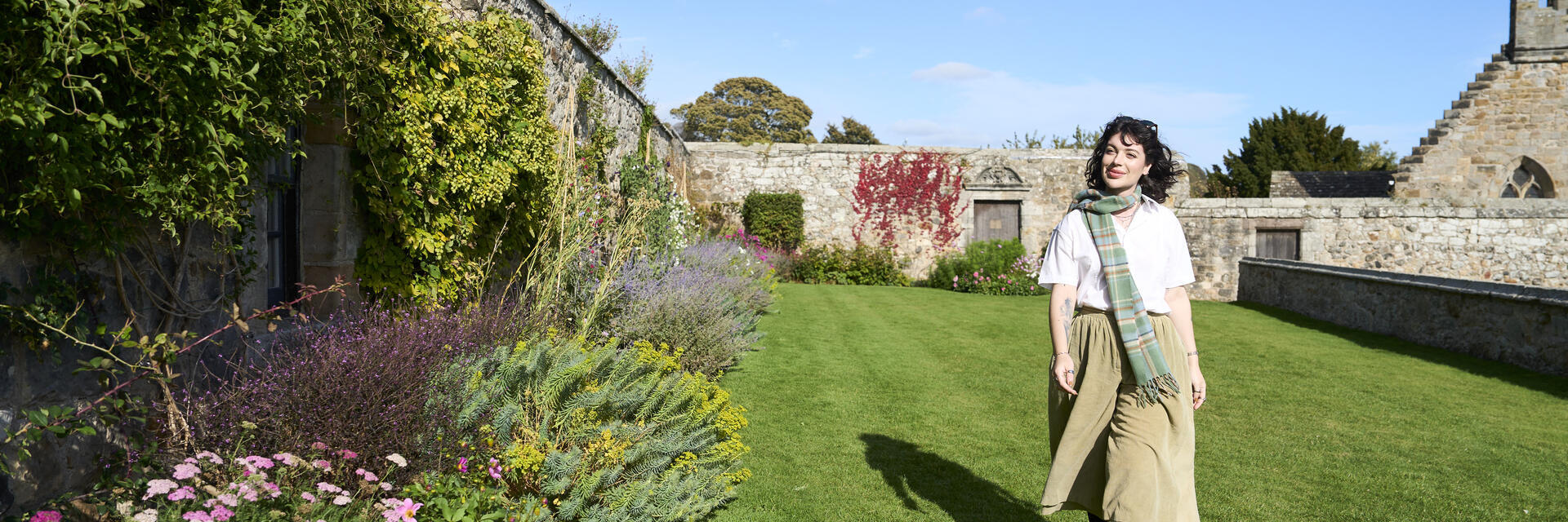 Frau geht im Garten neben einer Steinmauer, umgeben von Blumen und Pflanzen, unter blauem Himmel.