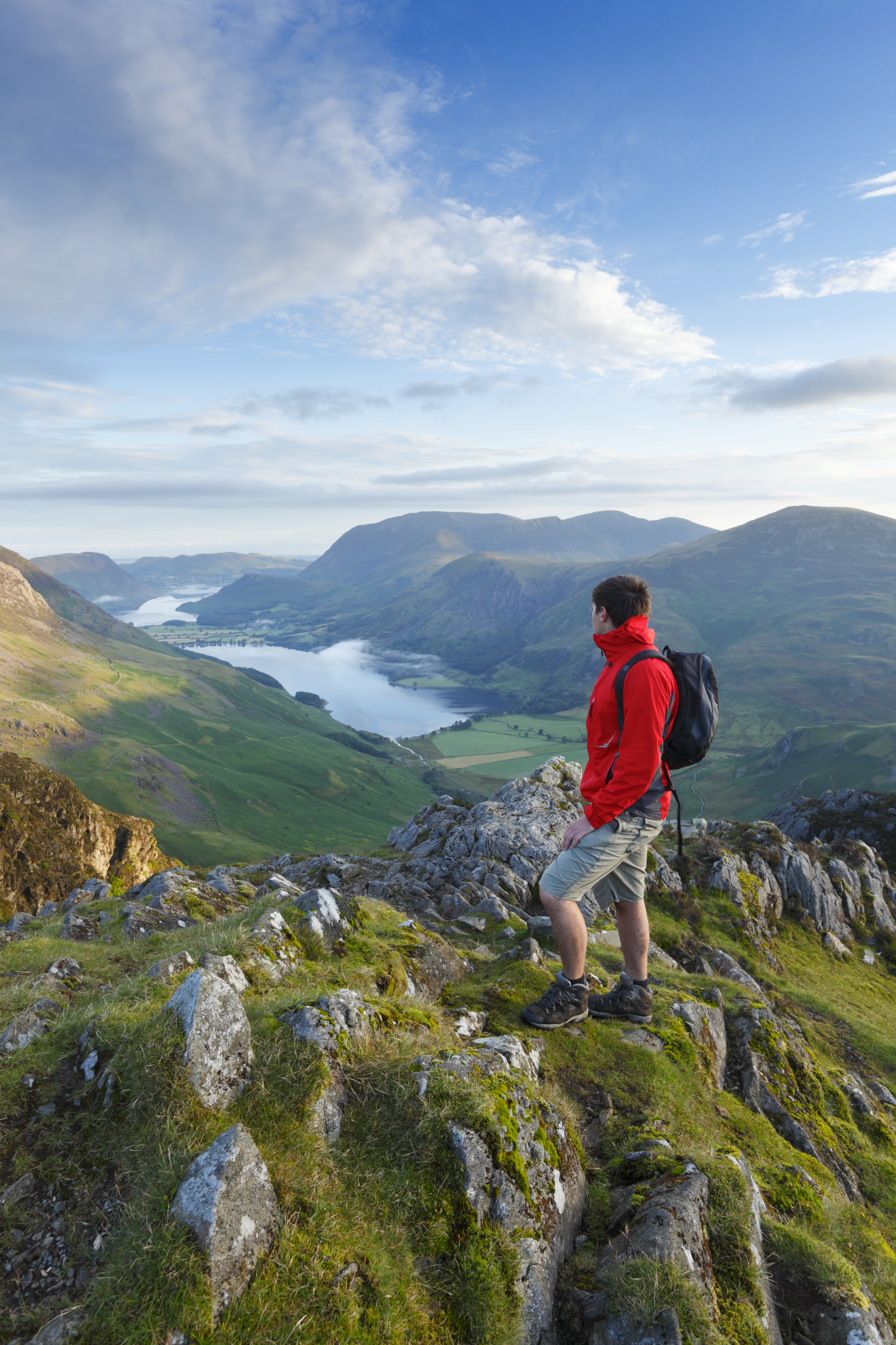 A man wearing a rucksack stands on a mountain fell overlooking hills and a lake