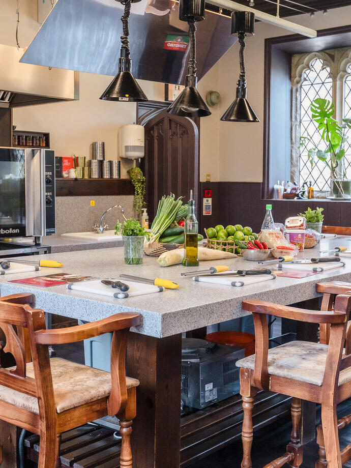 Modern kitchen with a central island, wooden chairs, prep stations, fresh ingredients, and a large oven. Bright window in the background.