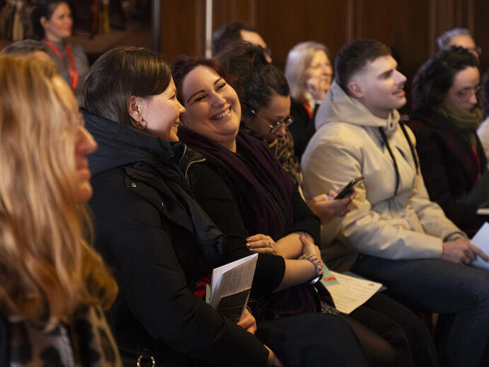 A group of people sitting indoors at an event, some smiling and laughing, with papers and phones in their hands.
