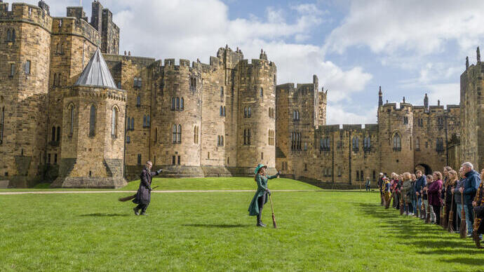 A group of people lined up for a Broomstick Training lesson with the wizarding professors by a large castle.