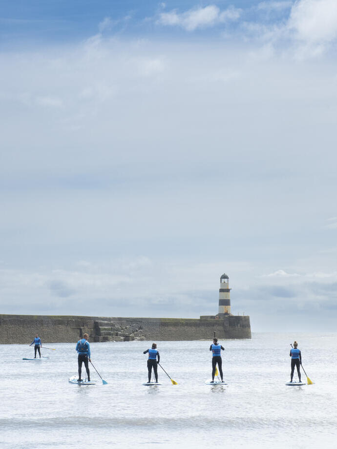 A group of people paddleboarding in front of a lighthouse and sea wall