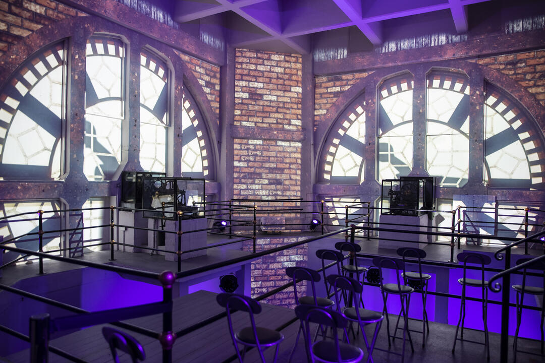 Interior with large clock face windows, exposed brick walls, metal railings, and rows of chairs lit by purple lighting.