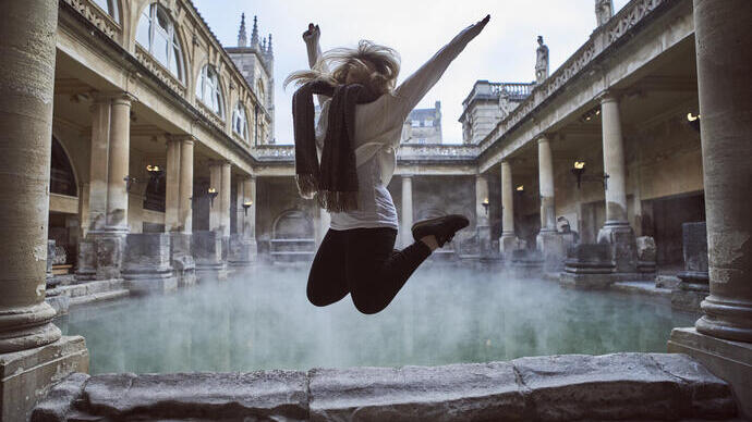 Frau springt in die Luft in einem römischen Freiluftbad, umgeben von historischen Gebäuden