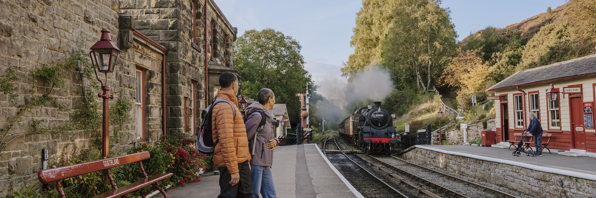 Gare rurale en pierre avec des voyageurs sur le quai et une locomotive à vapeur approchant. Arbres et bâtiments traditionnels autour.