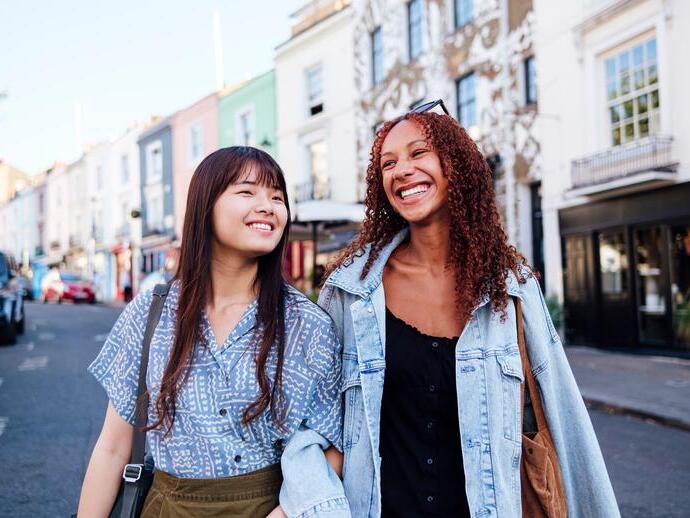 Two young women linking arms and walking in a street smiling