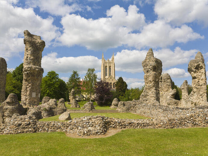 Ruinas de una abadía con una catedral al fondo, cielo azul con nubes y césped alrededor.
