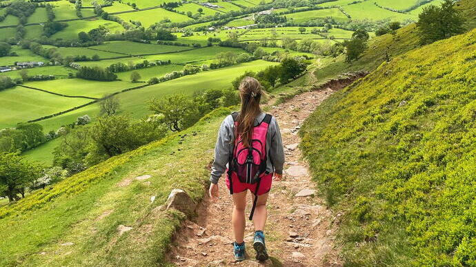 Rückansicht einer jungen Frau, die auf einem Weg durch ein üppig grünes Tal und Hügel spaziert