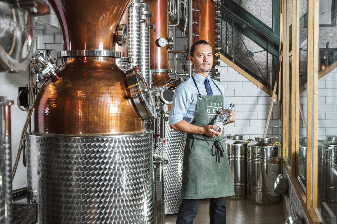 Uomo con grembiule verde in piedi accanto a un alambicco di rame in una distilleria di gin