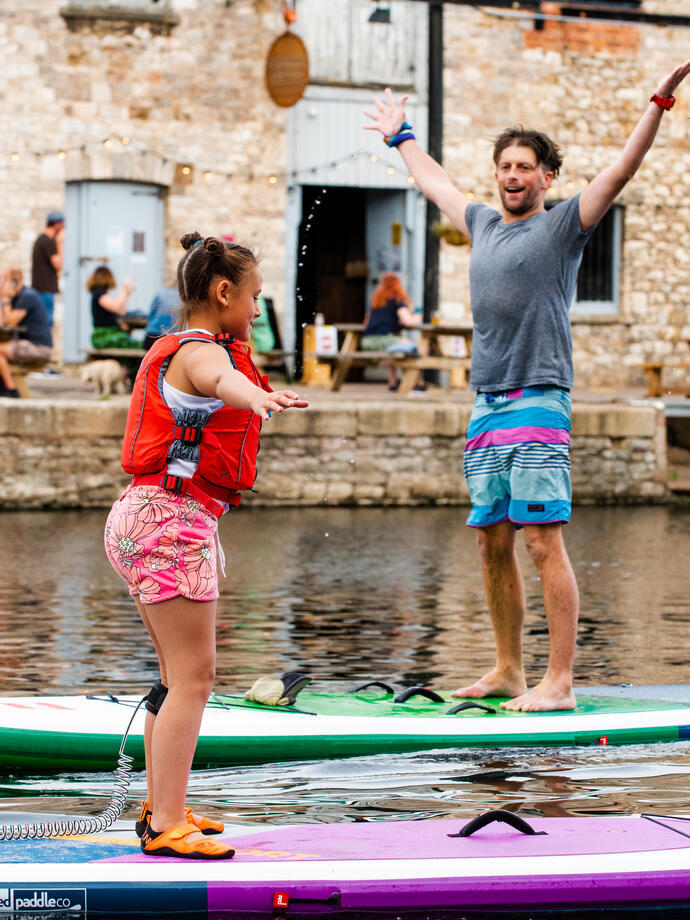 A child and a man stood on paddleboards in a river in an urban setting