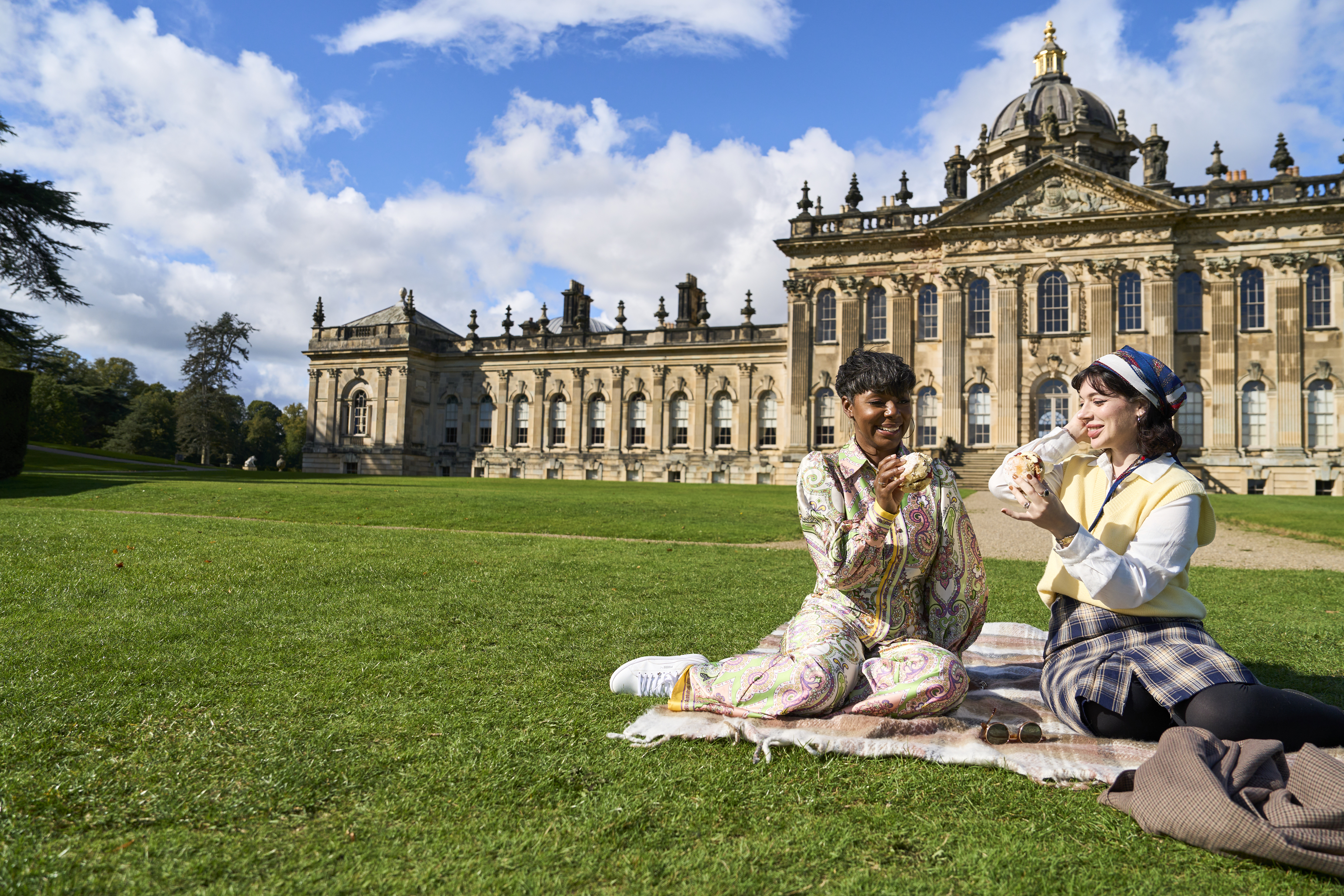 Zwei Personen sitzen auf einer Decke auf dem Rasen vor einem großen historischen Gebäude.