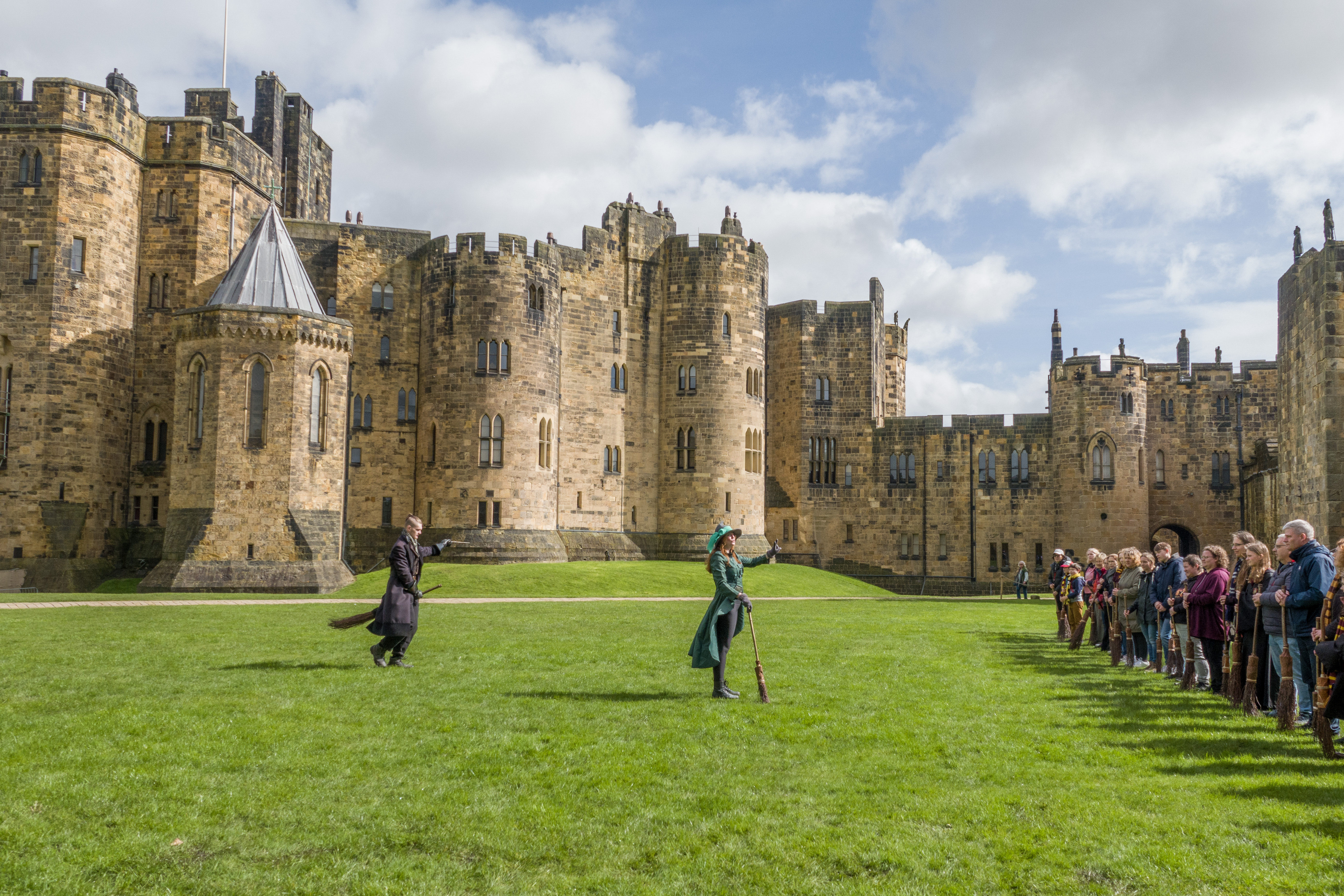 A group of people lined up for a Broomstick Training lesson with the wizarding professors by a large castle.