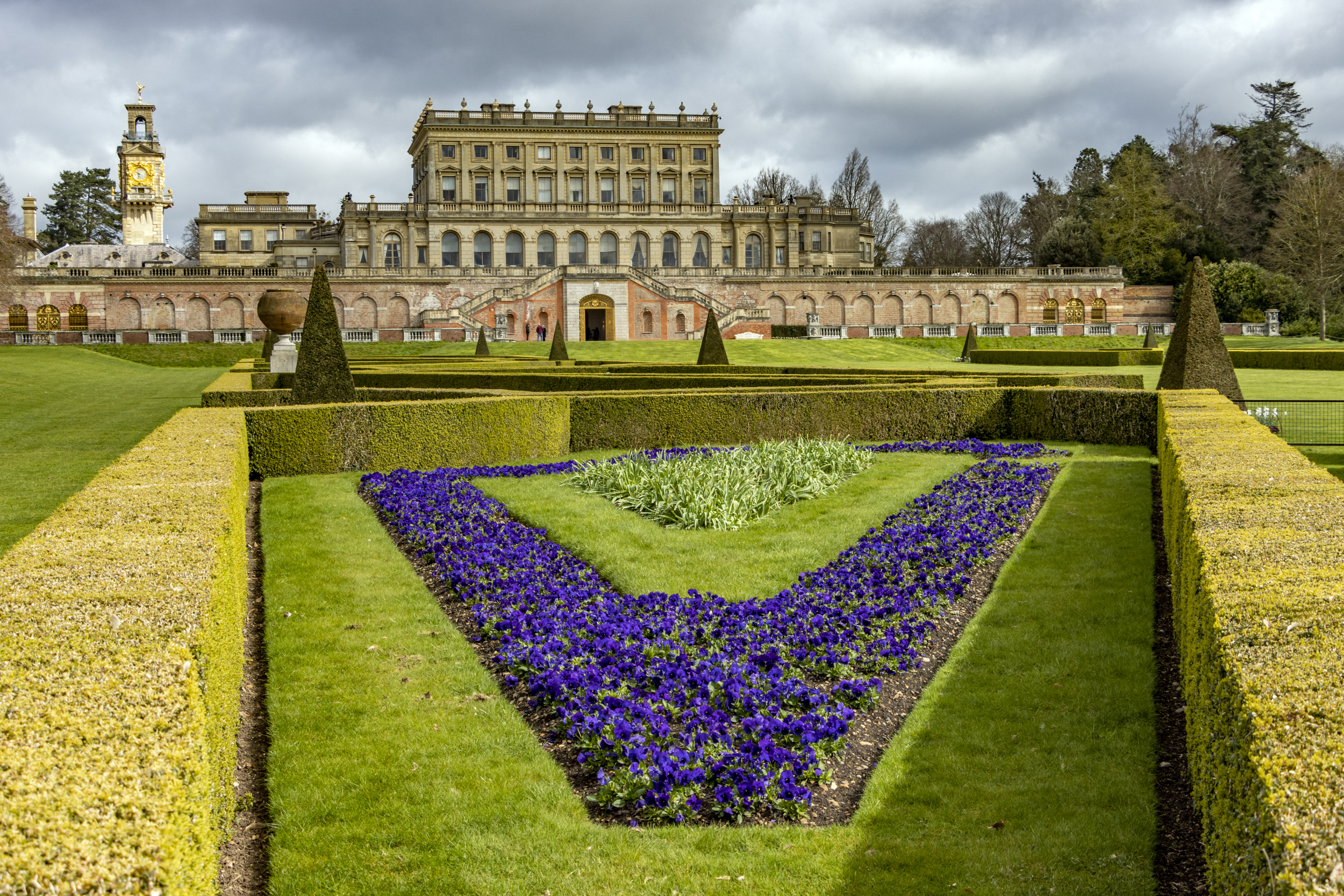 Beautiful manicured flower gardens and sculptured hedges in front of a large mansion.