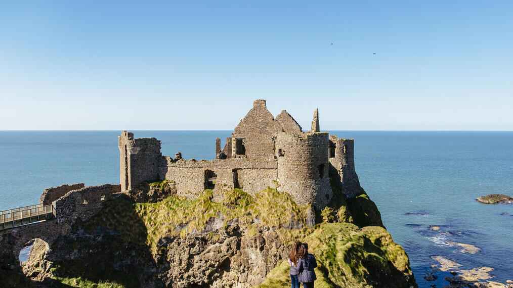 People looking at an historic stony castle from a rocky outcrop cliff over the ocean.